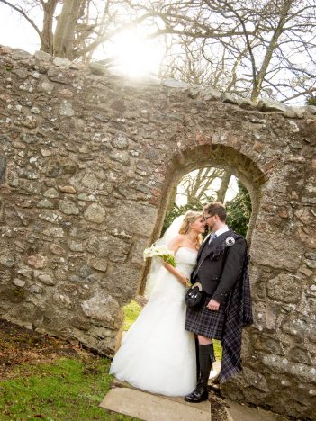 A bride and groom having their Maryculter House Hotel wedding photos in the grounds. The are standing below a stone arch