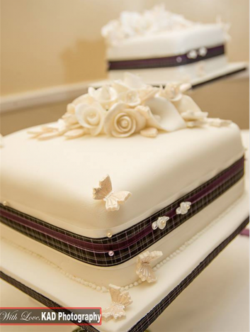 2 tiers of a wedding cake. There is a tartan and purple ribbon around the cake, and it has calla lillies, butterflies and roses on top
