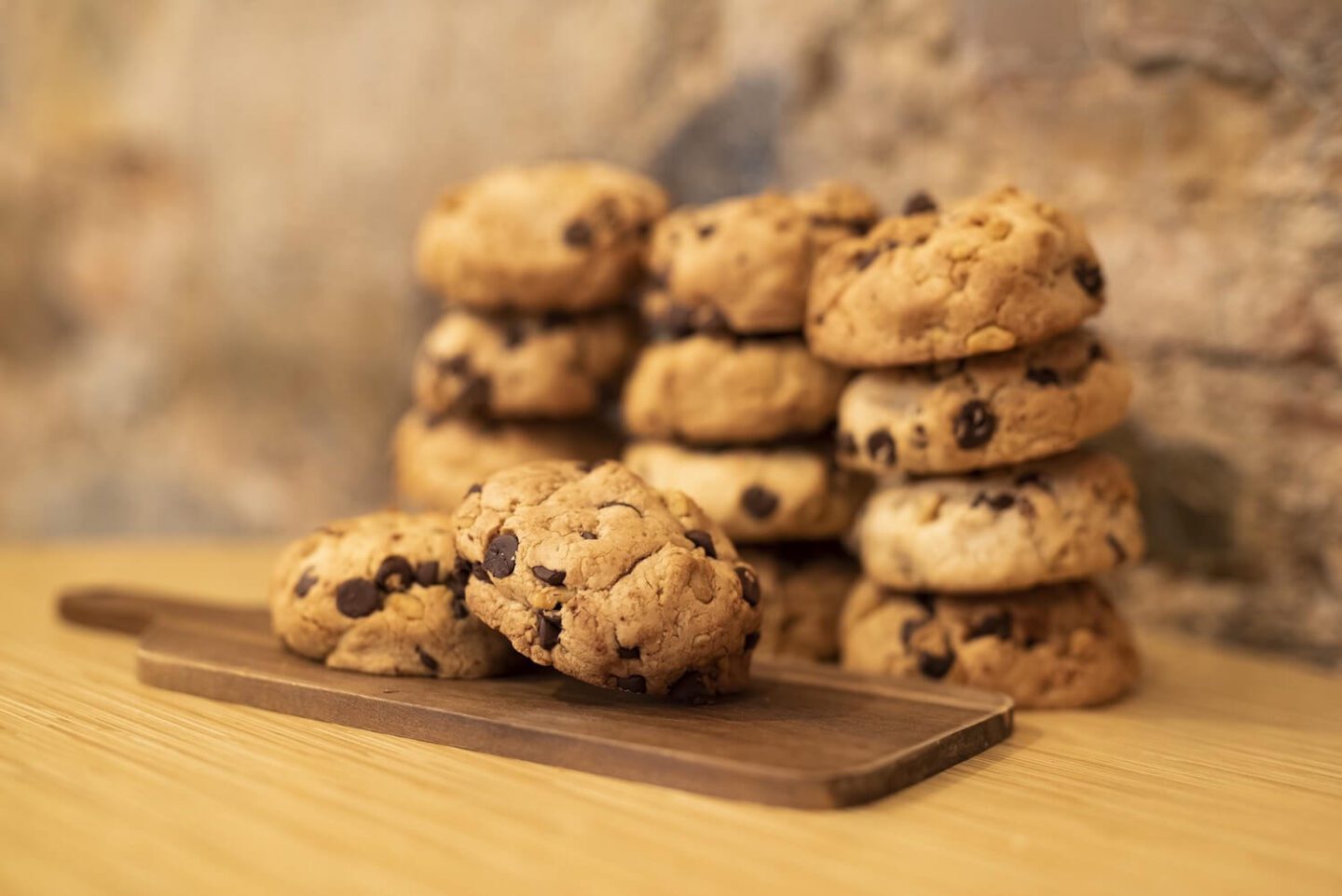 A wooden board with 2 chocolate chip cookies on it, and more cookies stacked behind