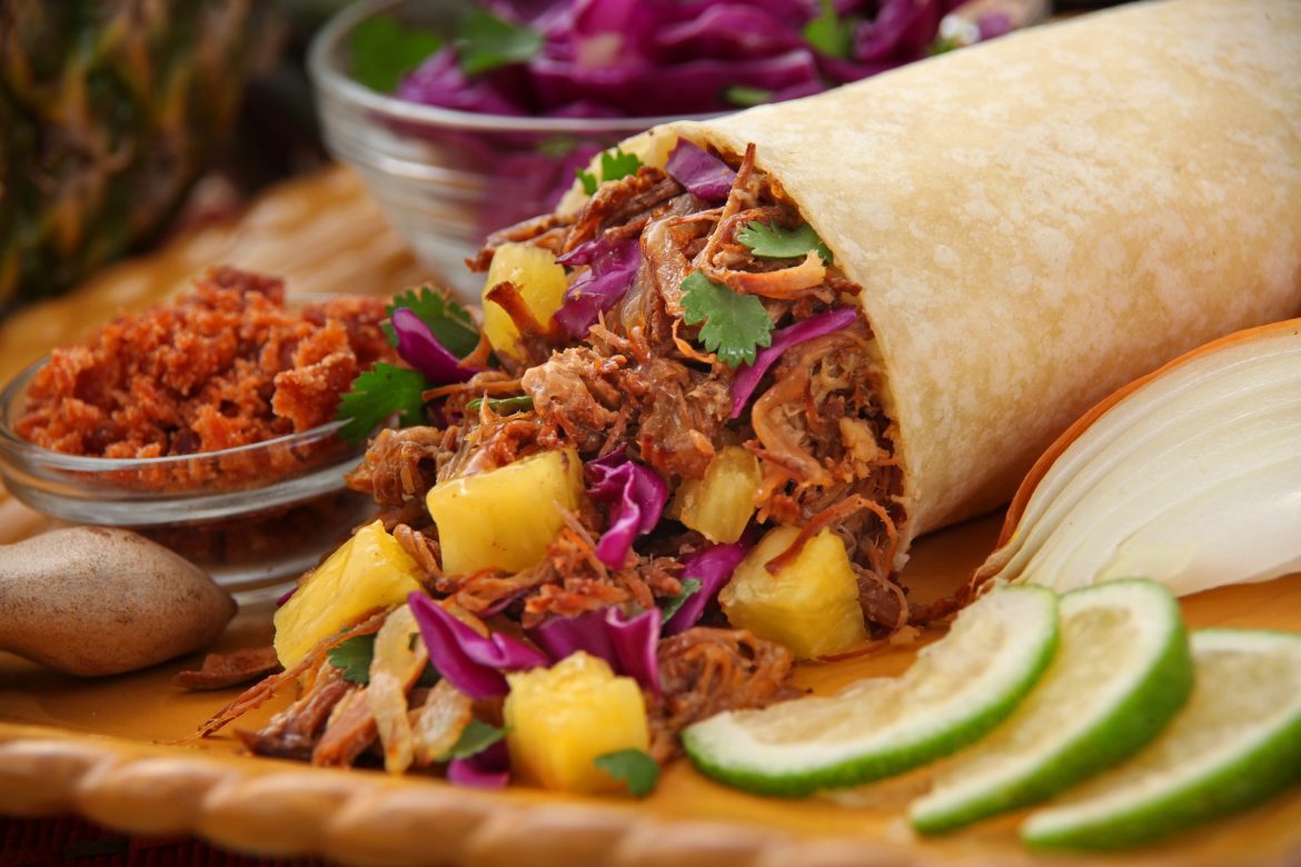 A pulled beef burrito with pineapple chunks, laid on a large board with lime wedges and condiments