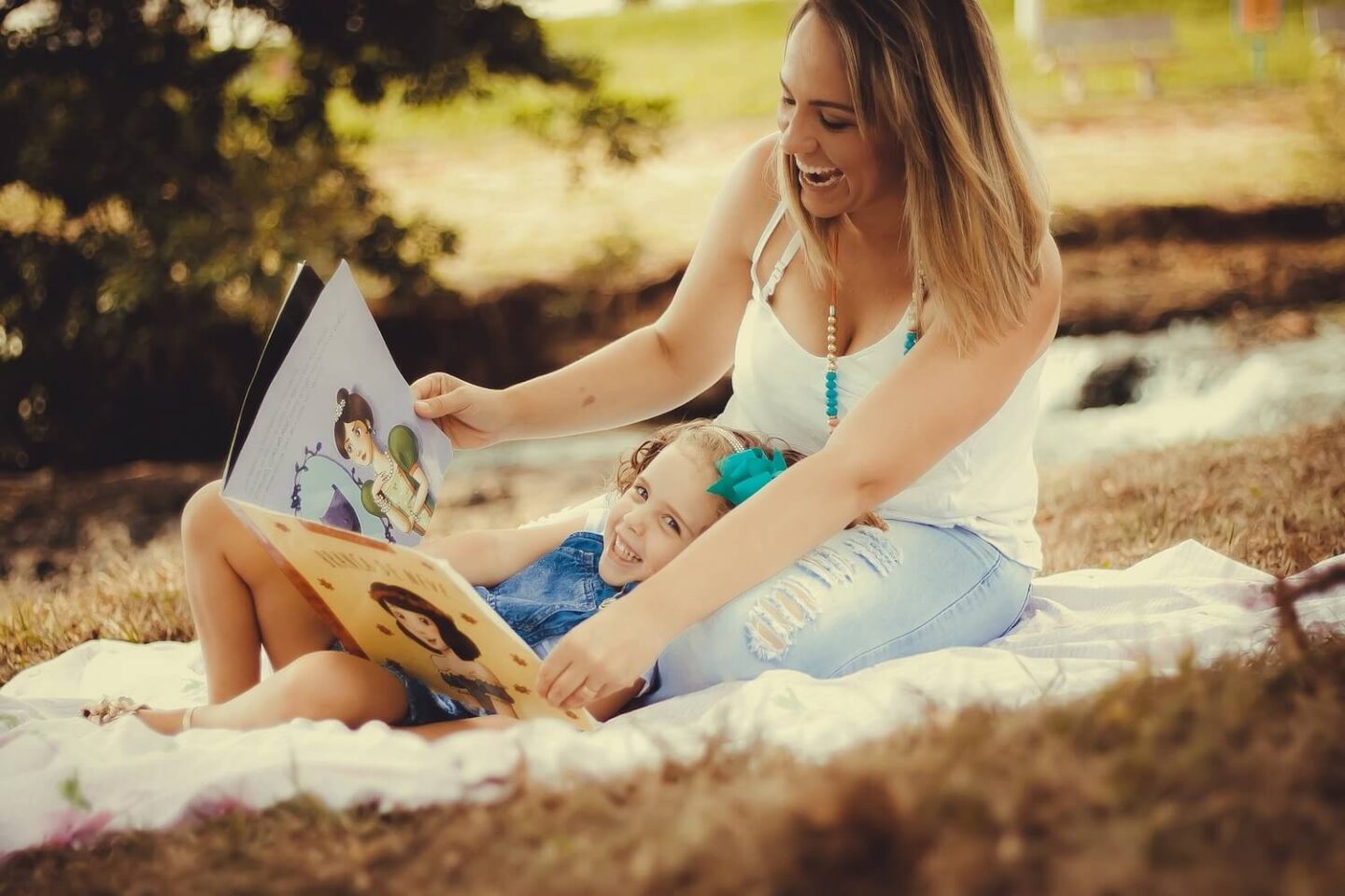 A woman sitting outside. A little girl is lying with her head in the womans lap and they are looking at a book