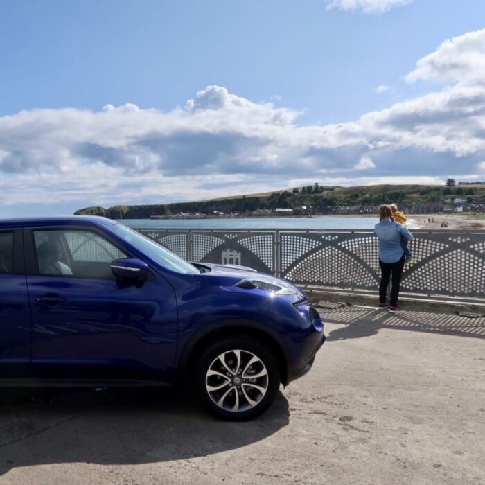 A blue Nissan Juke car parked next to the beach. A woman and child are in the background looking at the sea