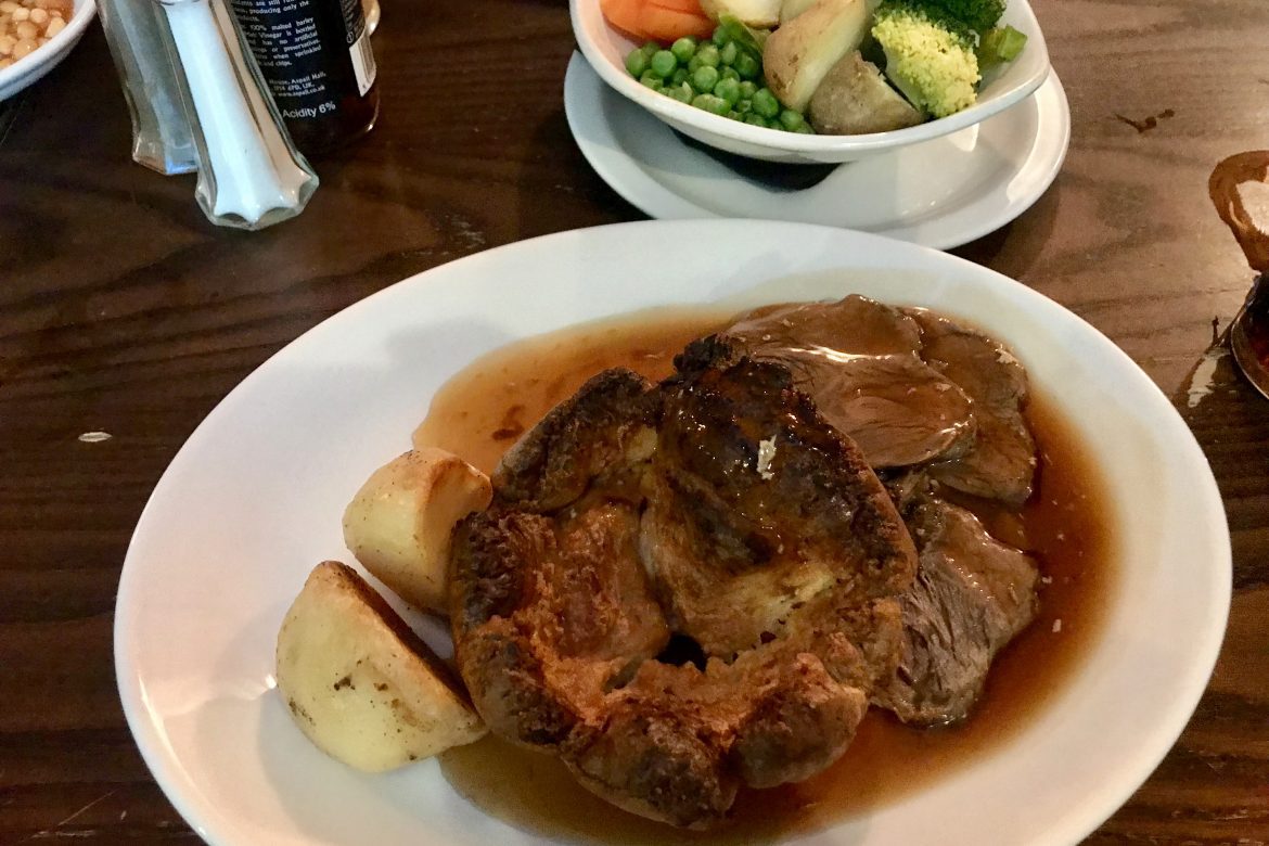 Roast beef, yorkshire pudding and roast potatoes with gravy on a place, with a dish of vegetables on a pub table