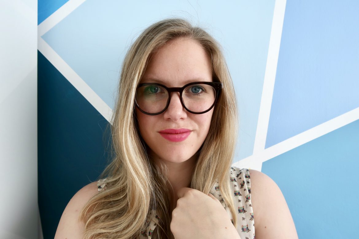 A woman with blonde hair, glasses and type 1 diabates is standing in front of a blue geometric wall
