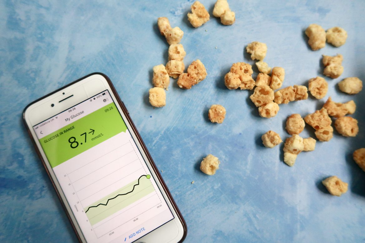 An iphone with a freestyle libre reading on it, next to a keto snack of puffed cheese
