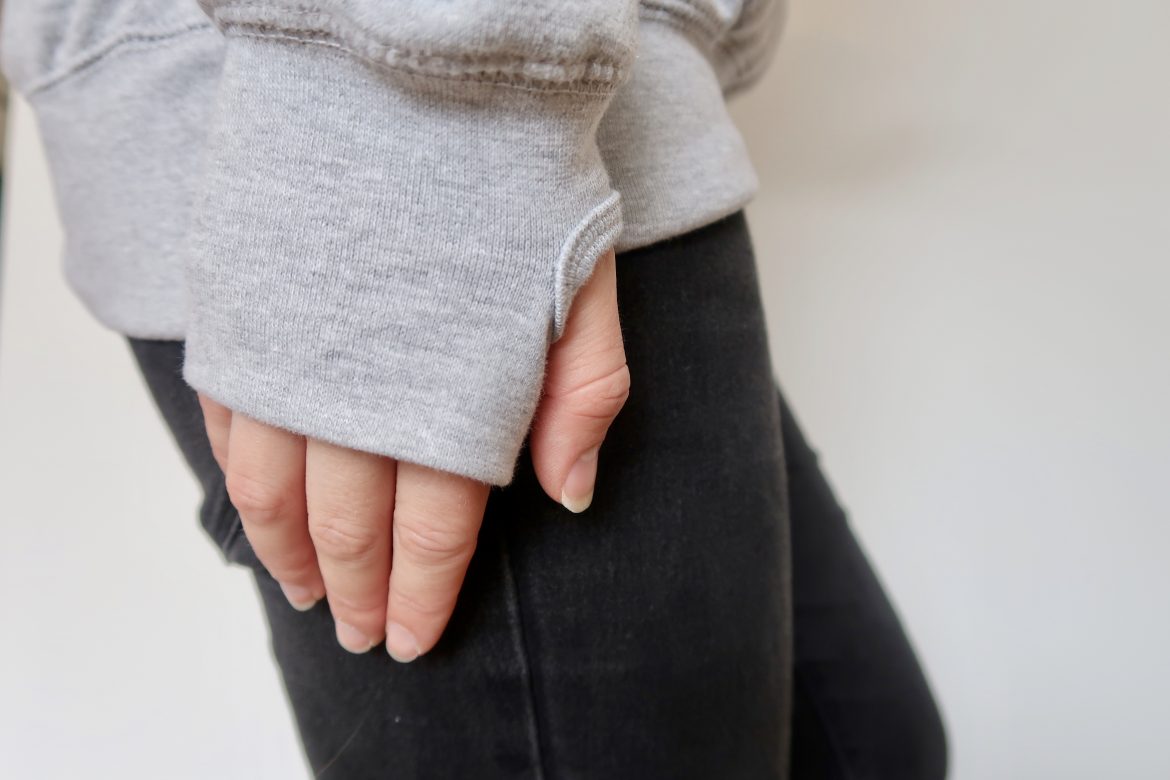 A hand in the sleeve of a grey hoodie, against a leg in black jeans