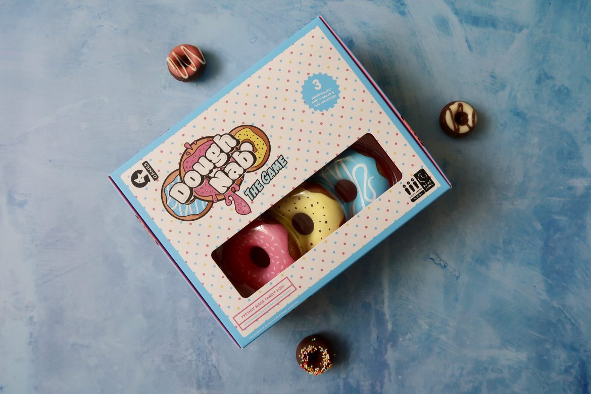 A game in a box called Dough Nab. Plastic donuts can been seen through the box, as well as 3 chocolate donuts surrounding the box.