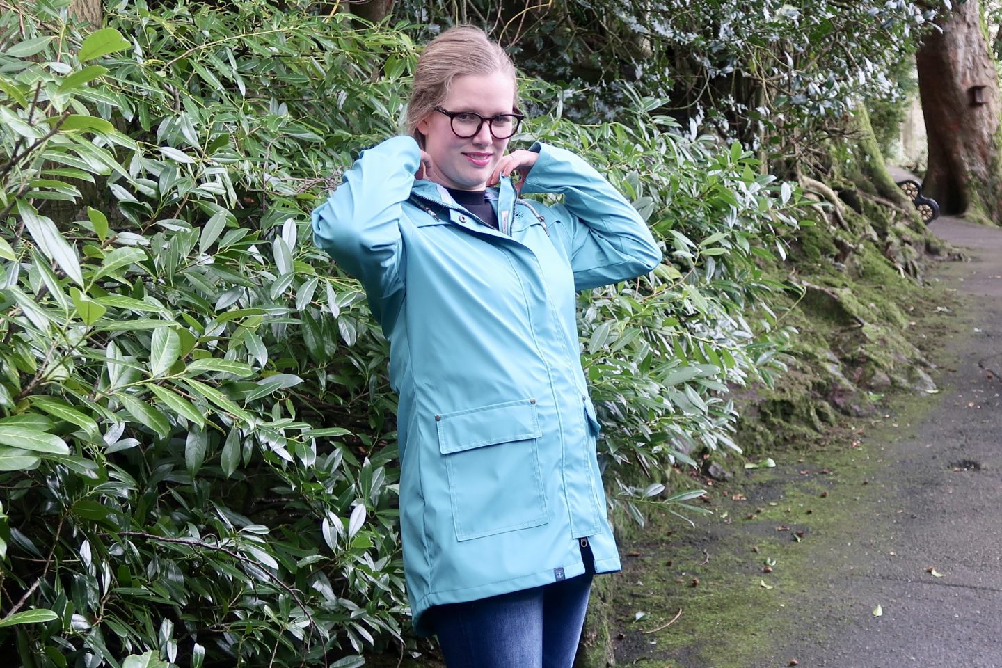 A woman in a teal jacket from Lighthouse Clothing. She has her arms up rearranging the hood on the coat