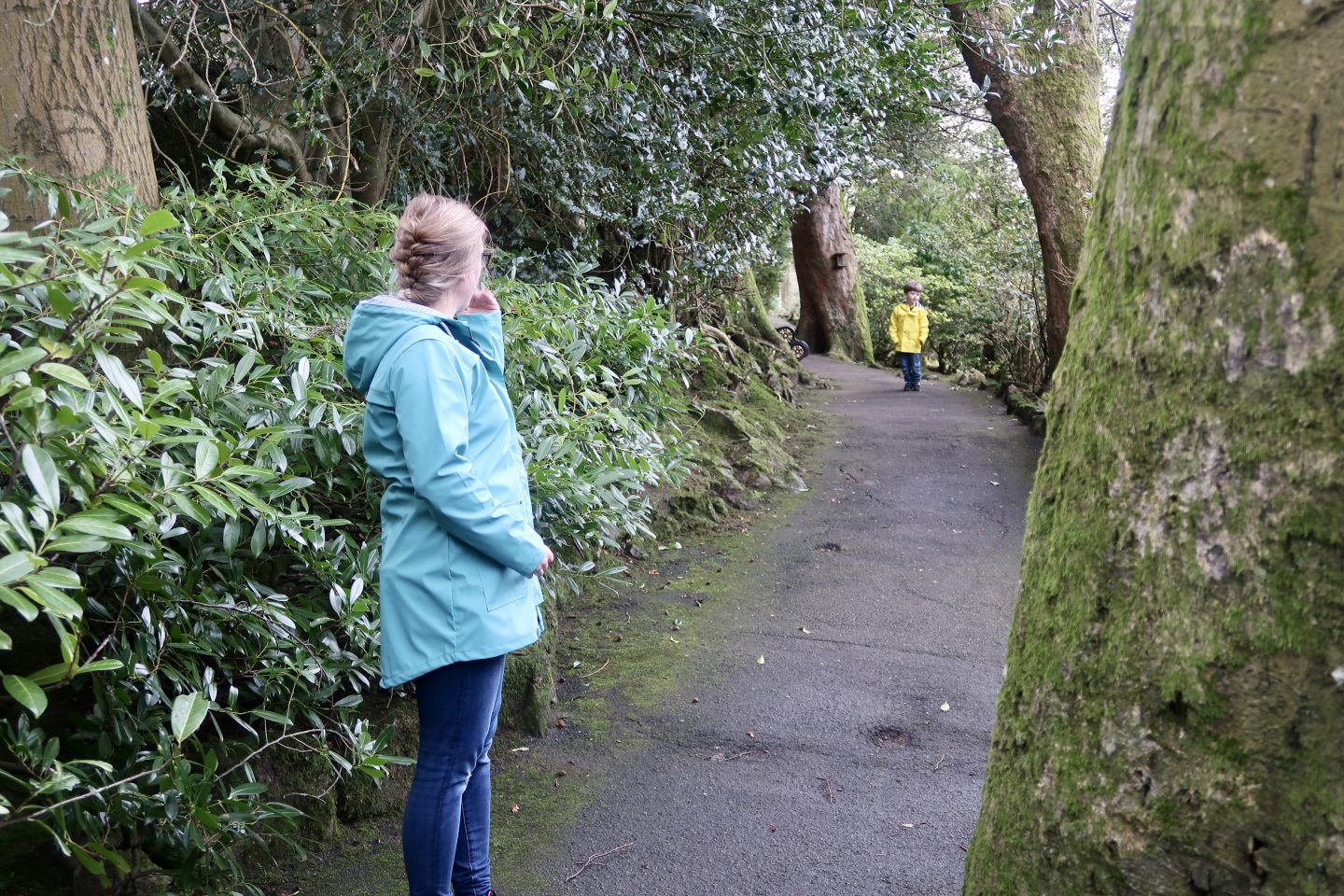 A woman standing in a teal jacket in a park, while a boy in a bright yellow jacket is in the background