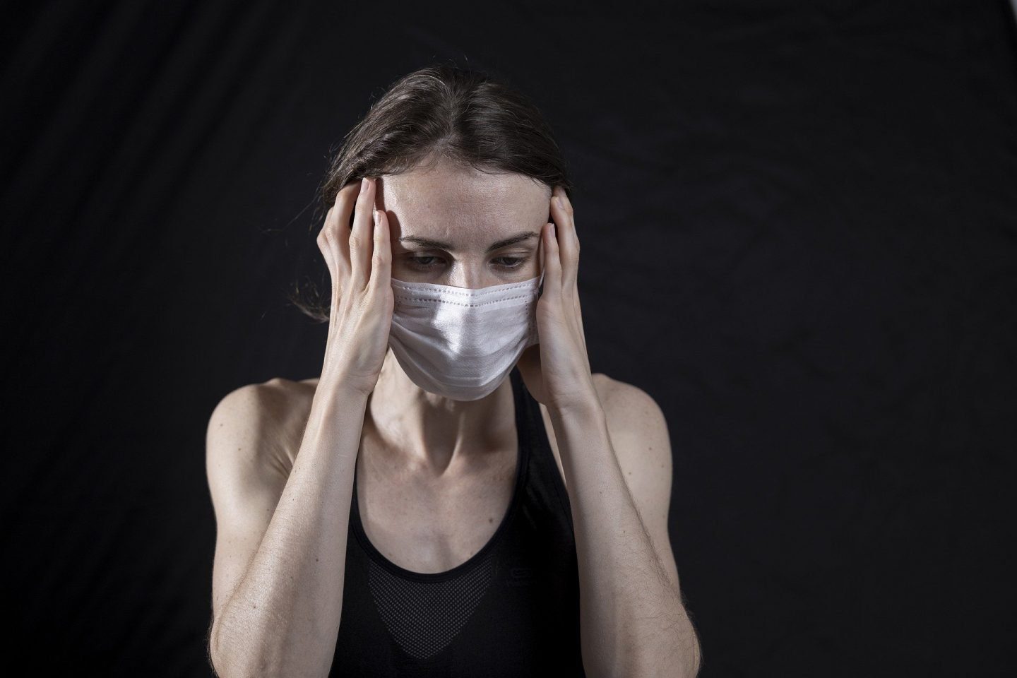 a woman wearing a black tank top and a white face mask. She has her hands up to her head as if she has a headache