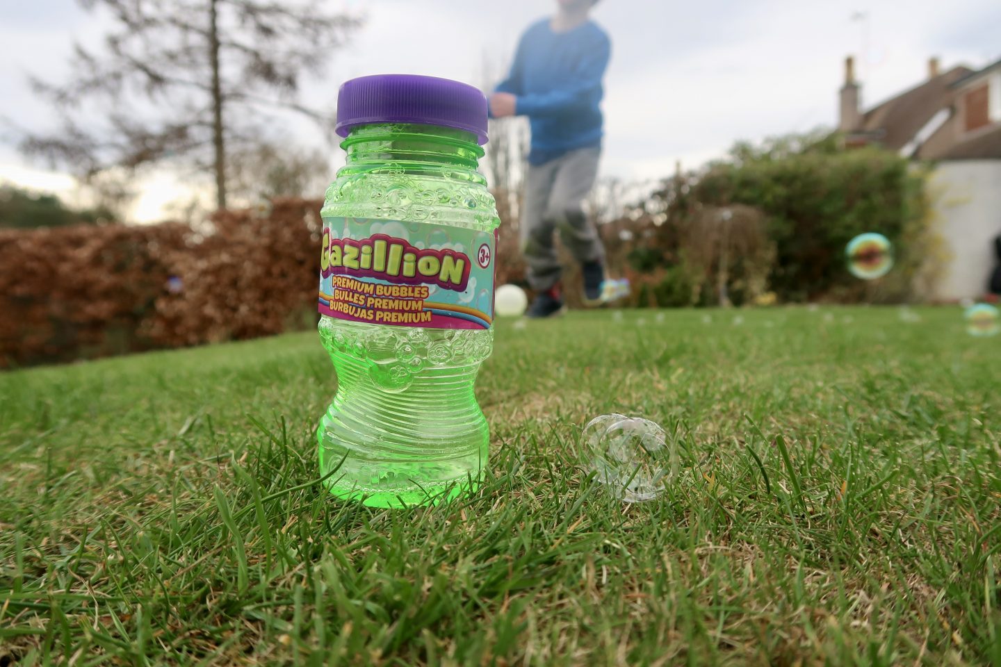 A bottle of bubble mixture on the grass next to a bubble, with a child playing in the background