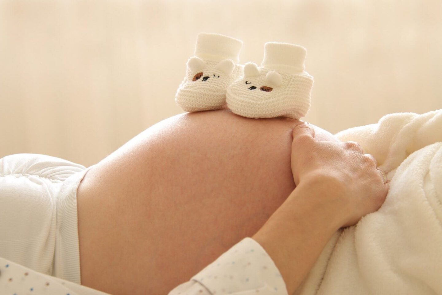 A pregnant woman lying down with a pair of knitted white boots on her bump