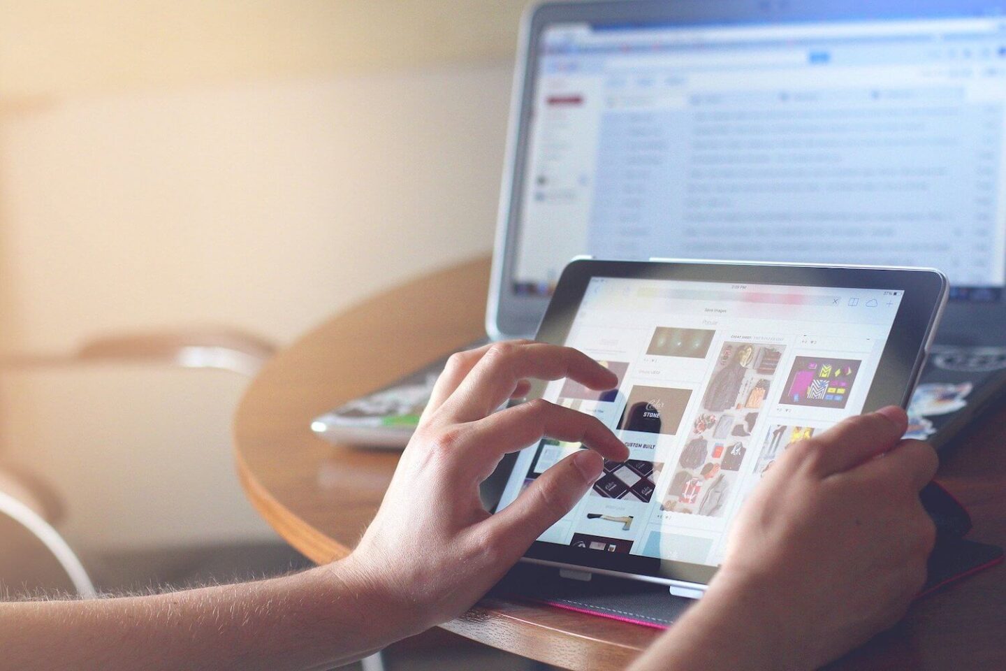 A hand touching the screen of an ipad with a laptop in the background