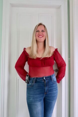 A woman wearing a red bodysuit and jeans. She has an insulin pump on her hip.