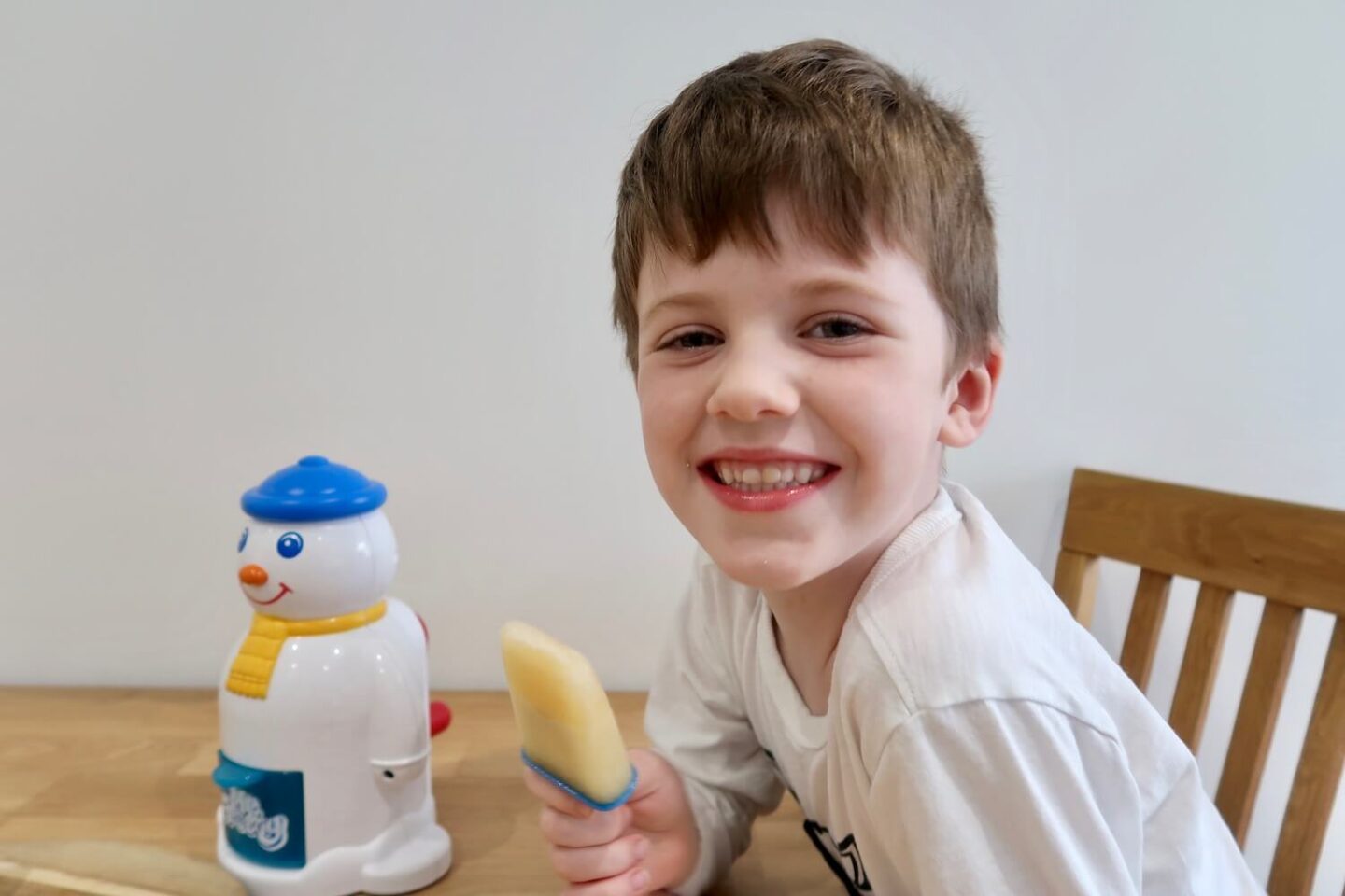 A boy smiling, holding a homemade ice lolly. he is sitting next to a Mr Frosty machine.