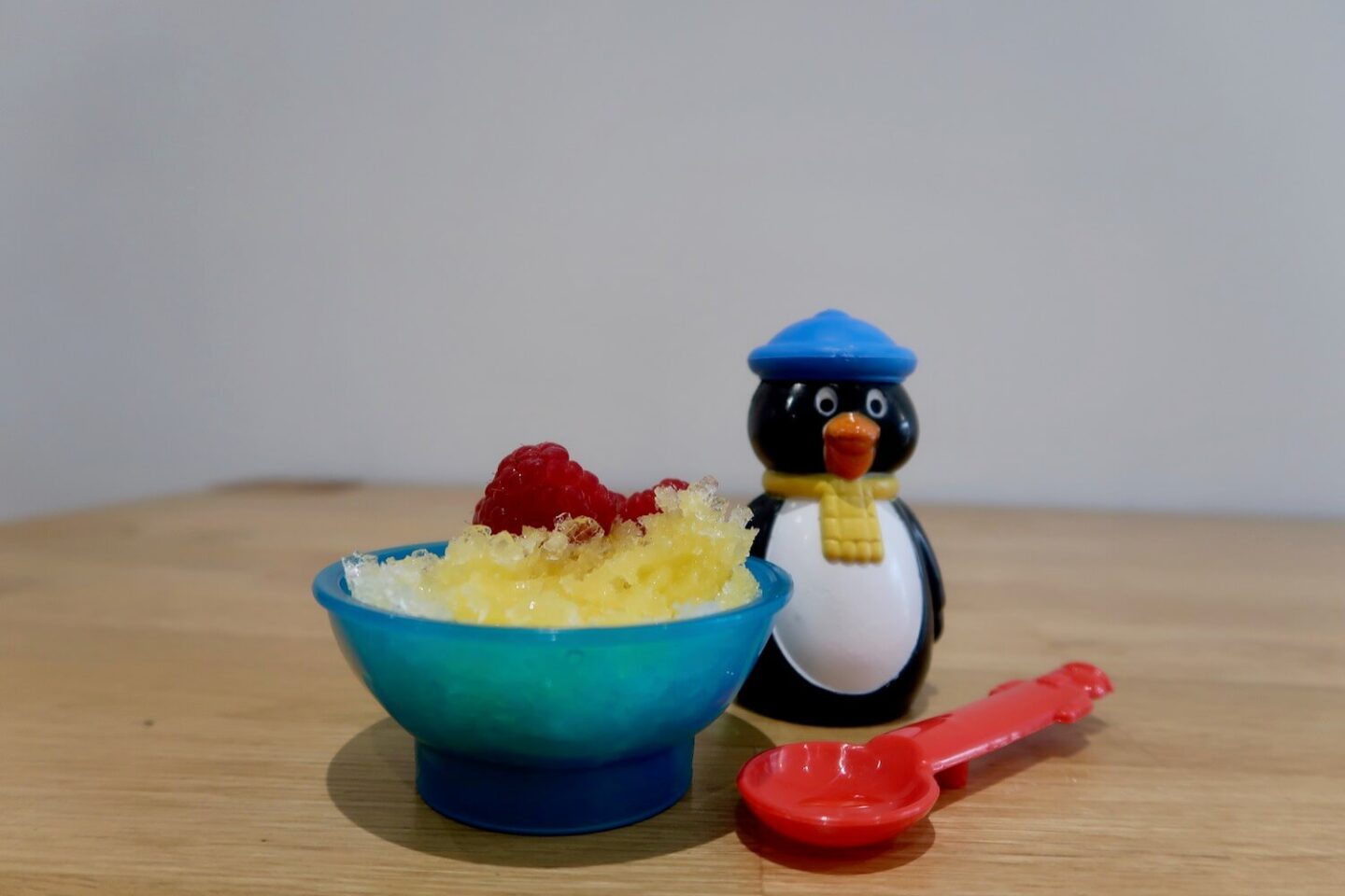 A bowl of orange crushed ice with some raspberries, next to a penguin and a red spoon
