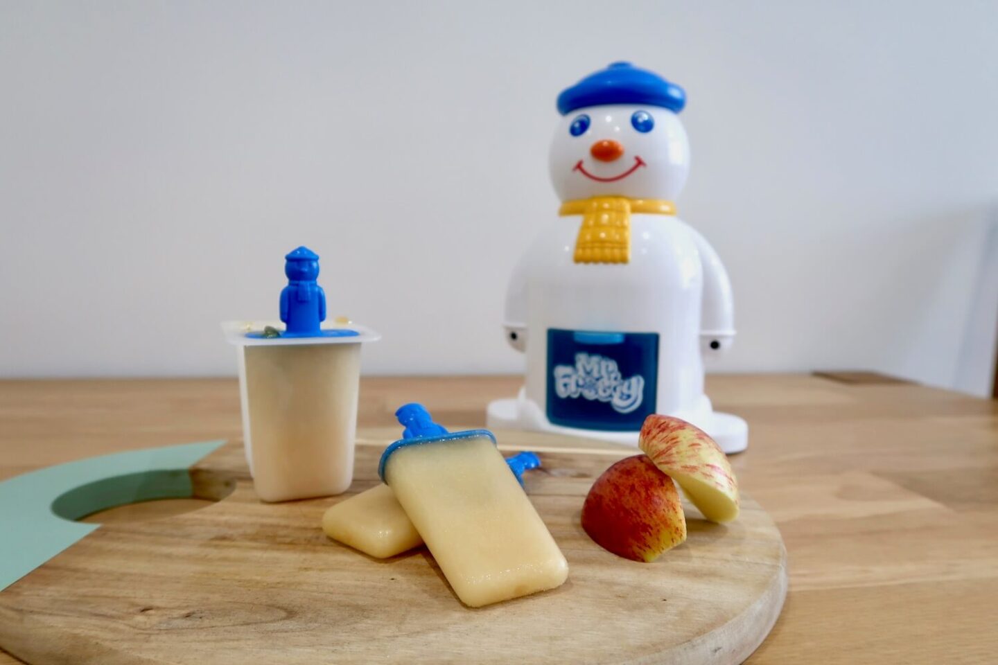 A wooden board with homemade apple ice lollies and some sliced apple, in front of a Mr Frosty machine.