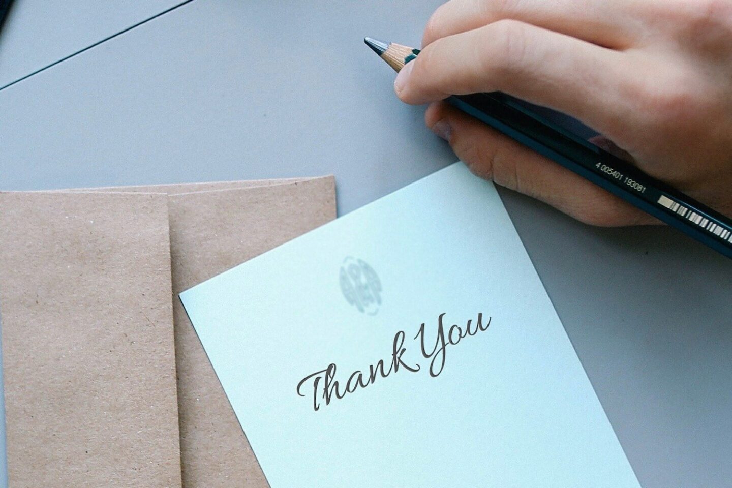 A hand holding a pen, next to a piece of note paper with "Thank you" written on it