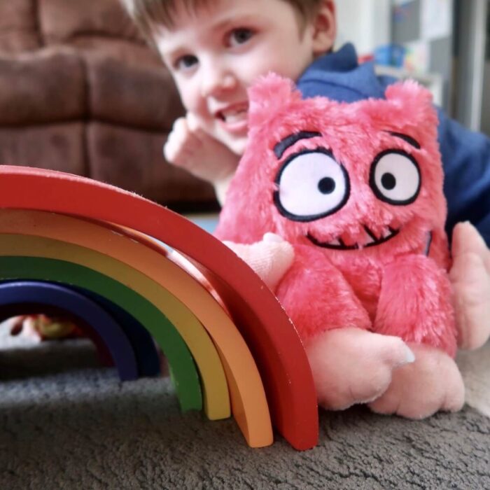 A wooden rainbow toy next to a pink fluffy monster you. A boy is smiling in the background