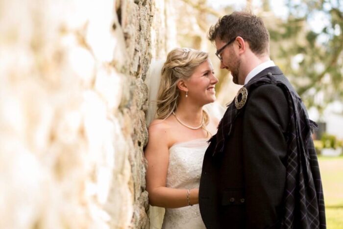 A blonde bride and dark haired groom lean against a wall and look at each other