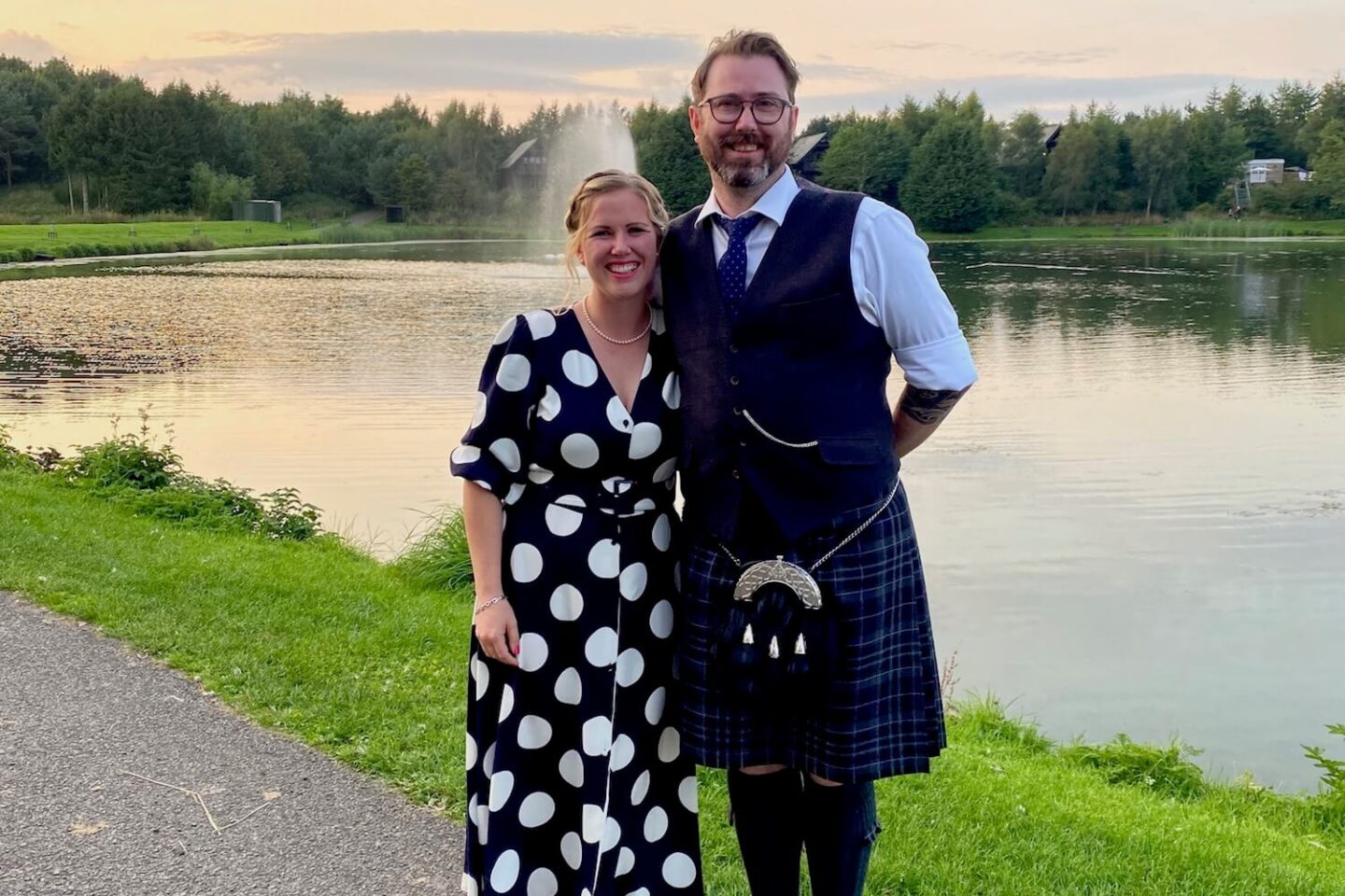 A man in a kilt and a woman in a blue and white spotty dress smile for the camera next to a lake