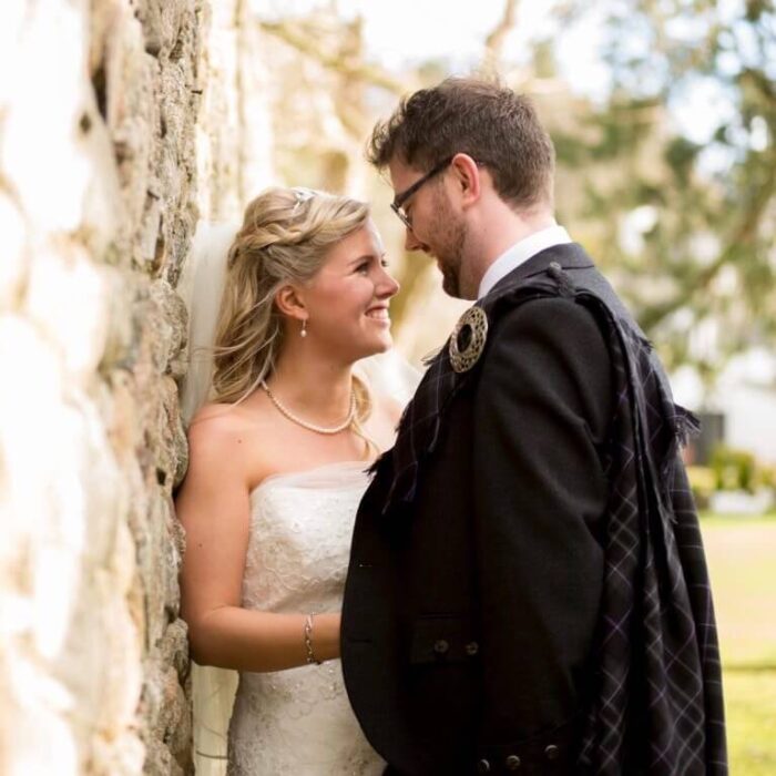 A blonde bride and dark haired groom lean against a wall and look at each other