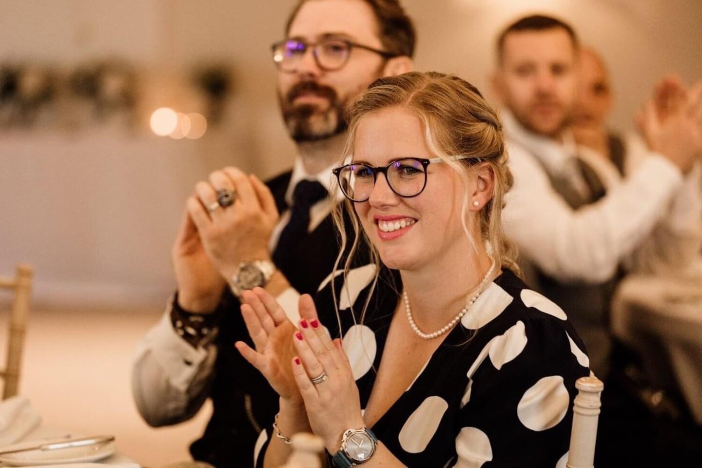 A woman in a blue spotted dress sat at a table at a wedding and claps