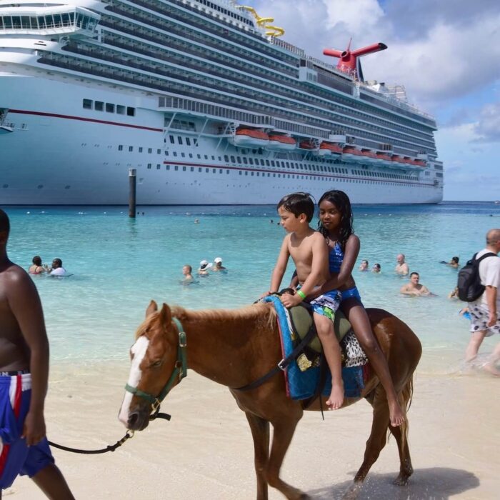 2 children riding on a horse on a beach with a cruise ship in the background.