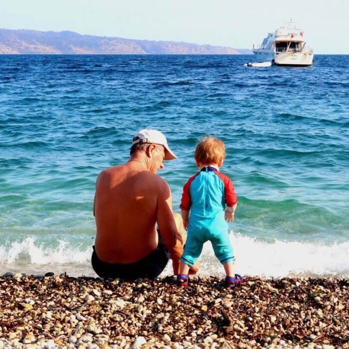 A toddler and his grandad on the beach in Corfu. The beach is stoney and the sea is bright blue