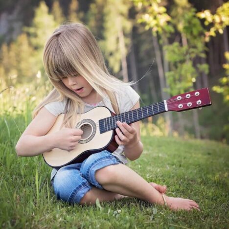 A blonde girl sitting on the grass plating a small guitar