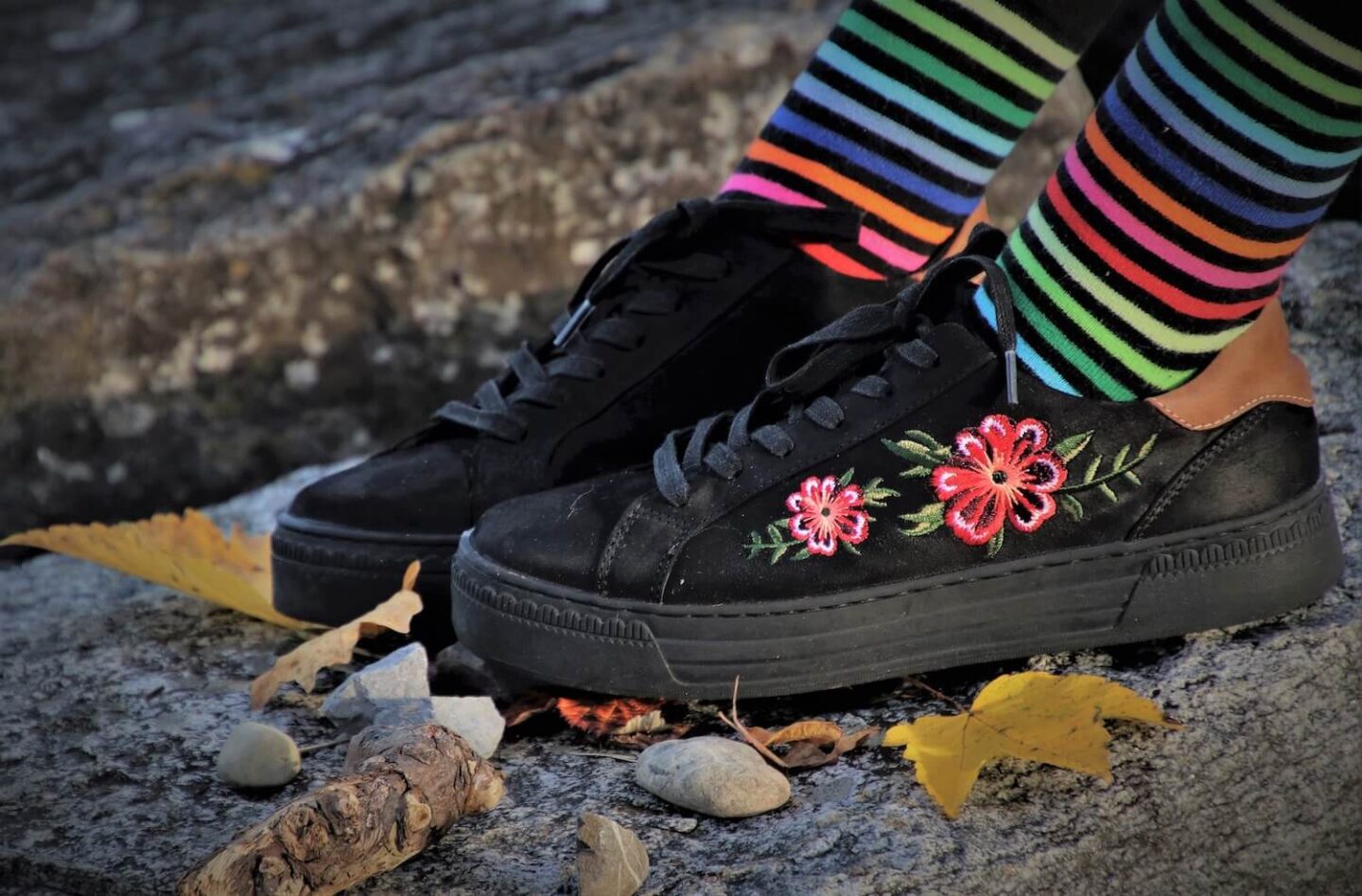 Black shoes with pink flowers embroidered on them, with rainbow striped socks on the persons legs