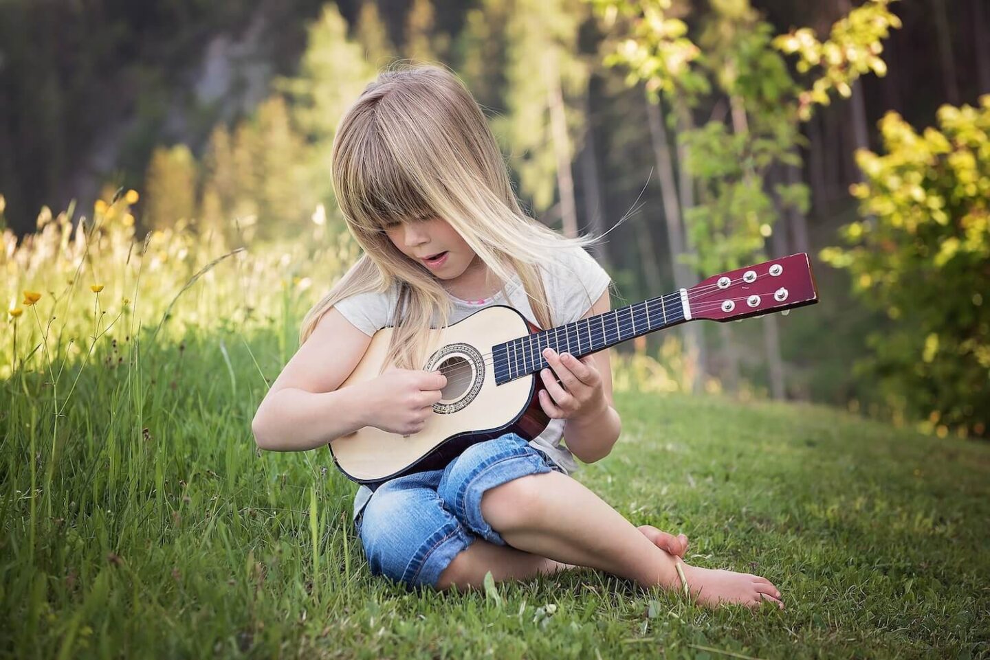 A blonde girl sitting on the grass plating a small guitar