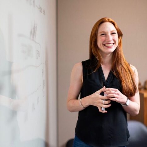 A woman with long red hair is smiling and standing next to a whiteboard
