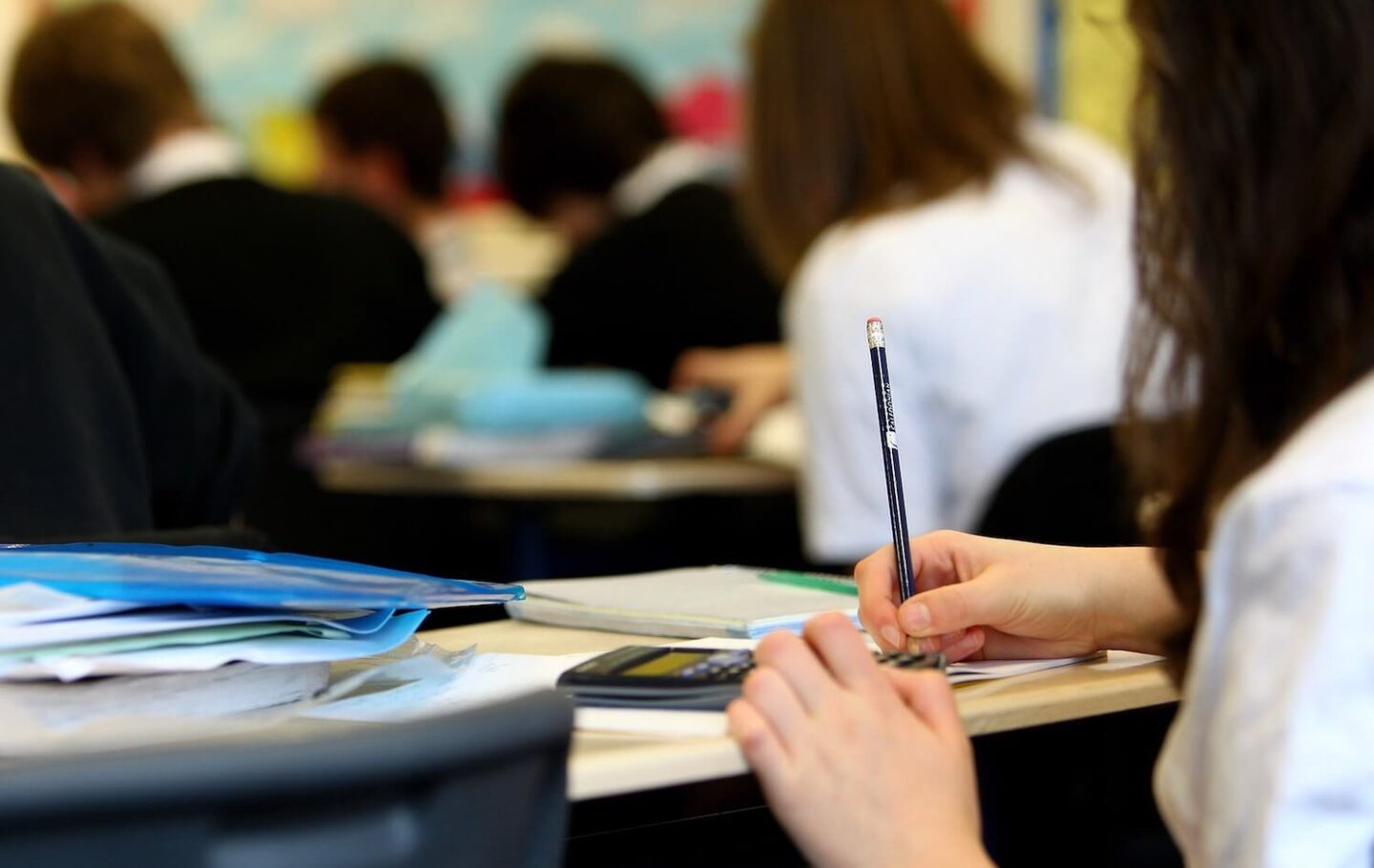 A desk of a child at school. Hands are holding a pencil and a calculator is visible.