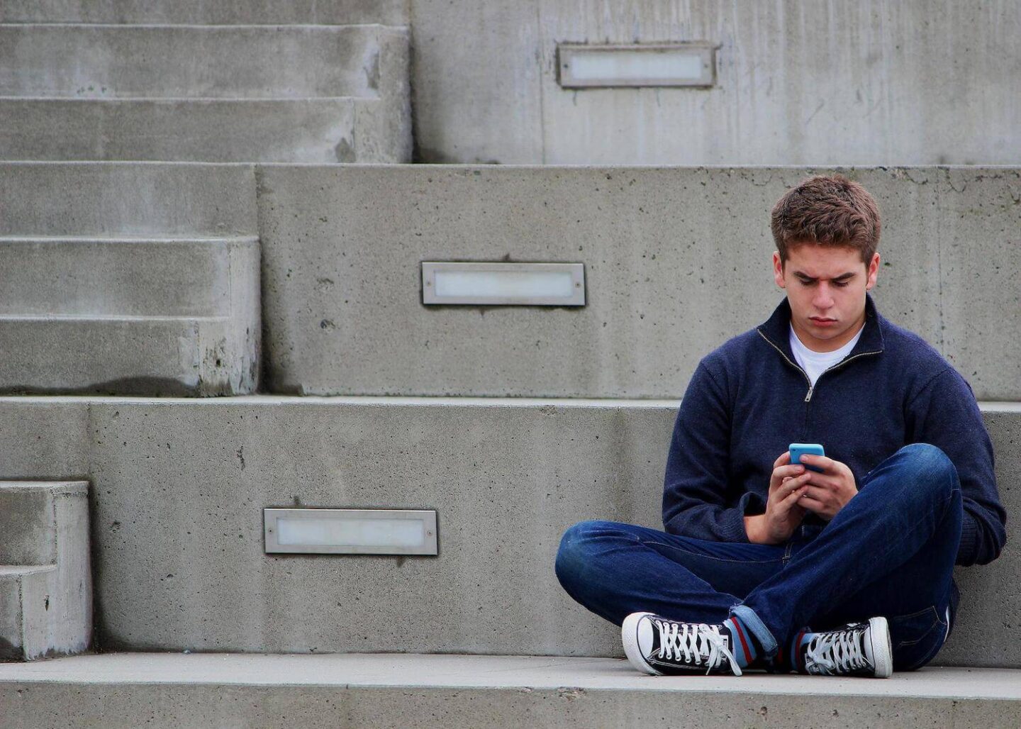 A teenage boy sitting on some concrete steps. He is looking at his phone.