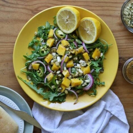 A yellow plate on a wooden table. It is filled with a salad and a lemon cut in half.