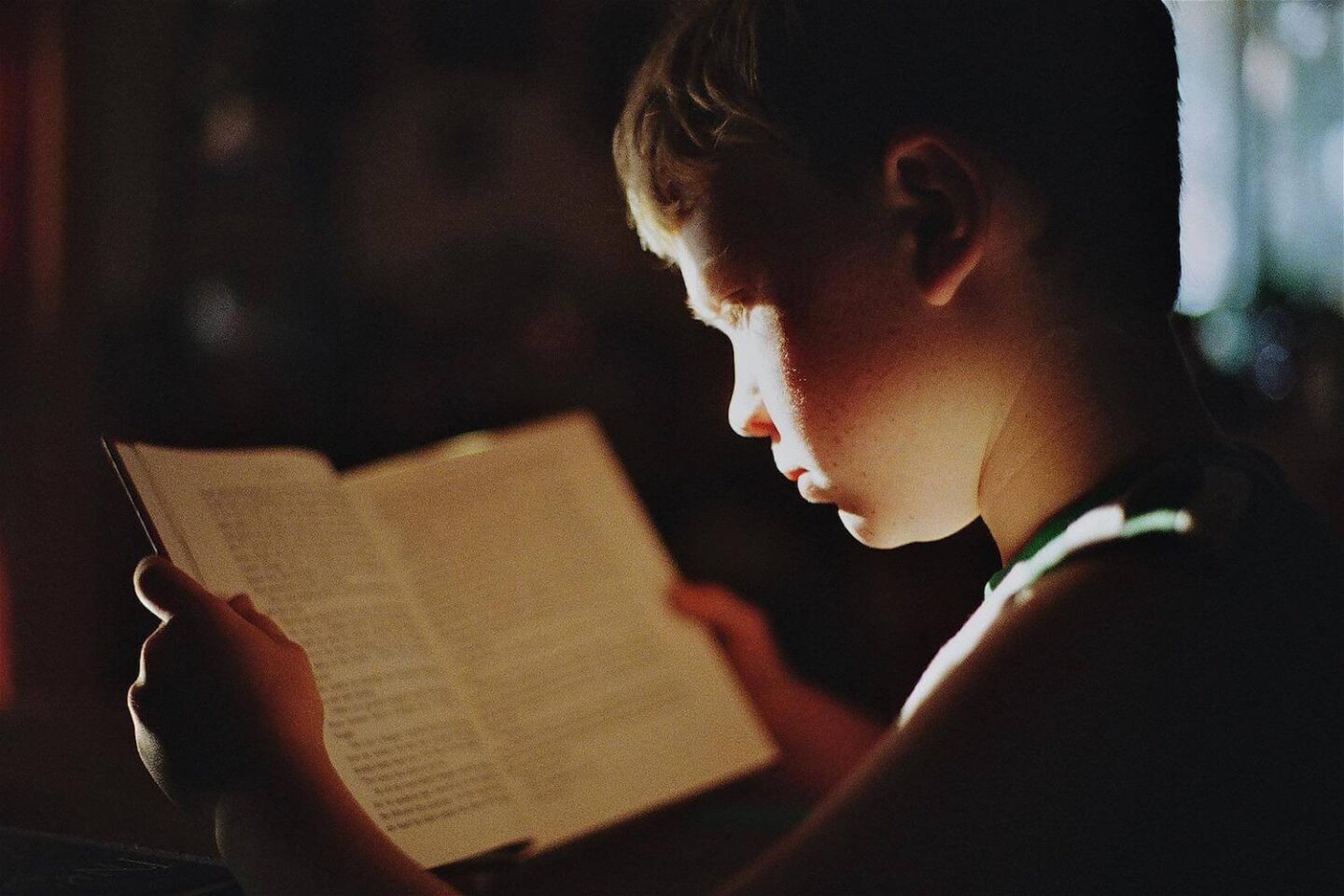 A boy reading a book with light shining on the side of his face.