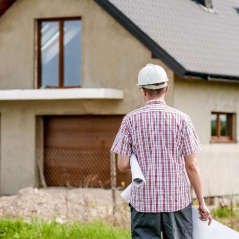 an architect walking towards a house. He has a checked shirt on and a white hard hat and it carrying some rolled up drawings