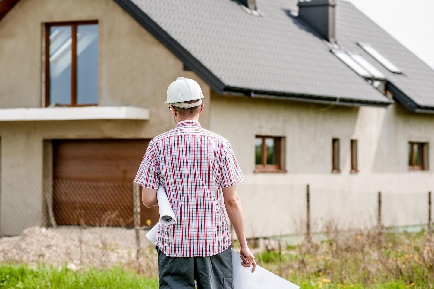 an architect walking towards a house. He has a checked shirt on and a white hard hat and it carrying some rolled up drawings