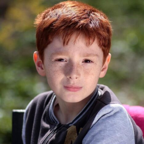 A boy with dark ginger hair and freckles looking at the camera with a serious expression on his face