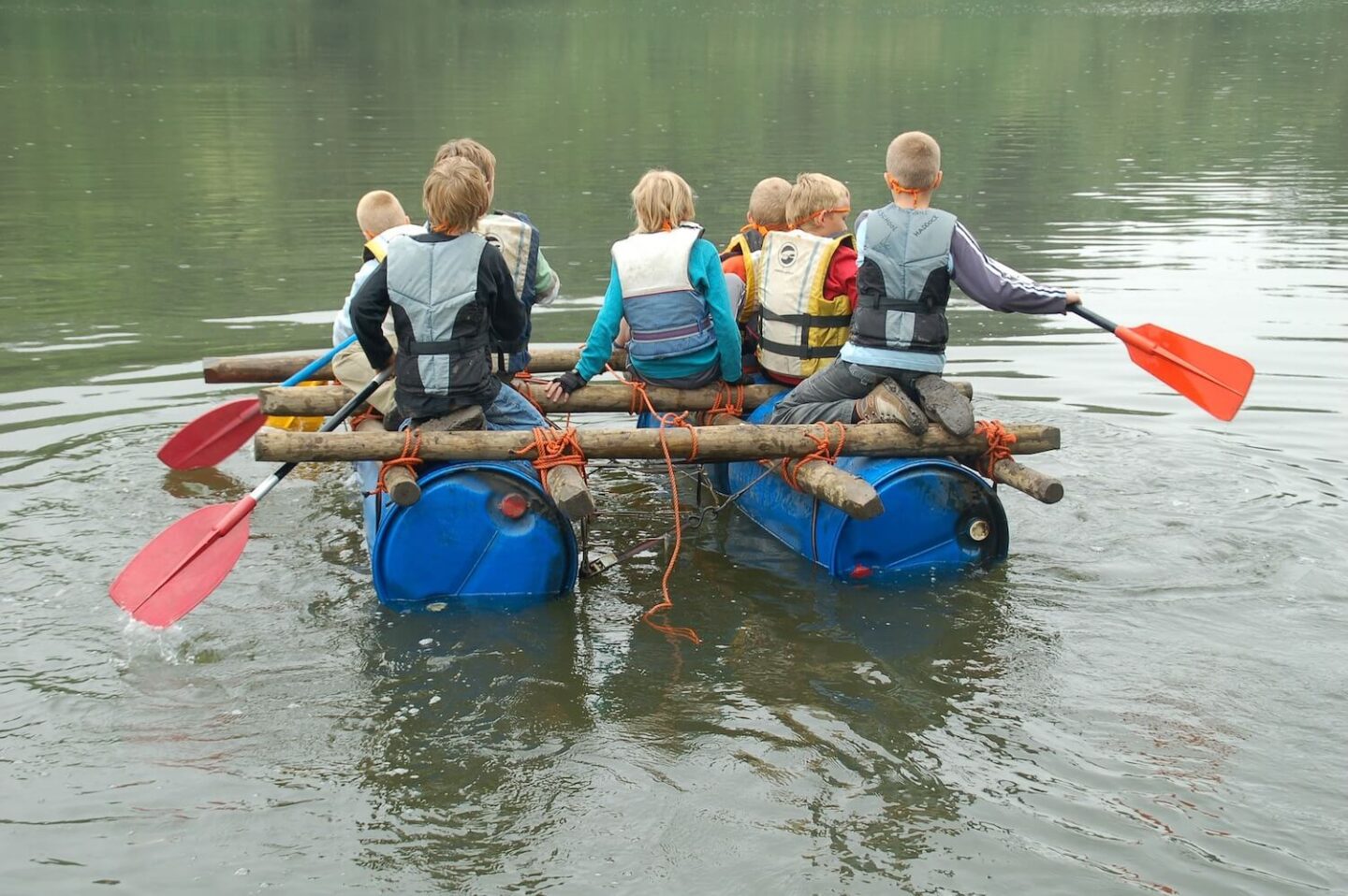 A group of school aged children rowing a homemade raft. It is made out of barrels, logs and rope