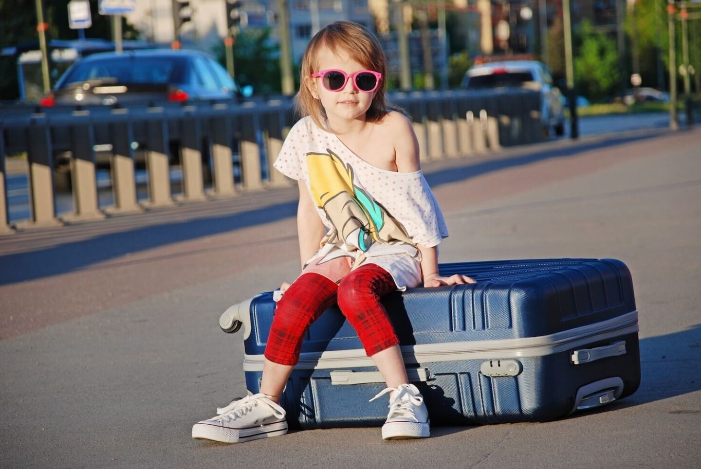 A young girl sitting on top of a suitcase in the middle of the street. She is wearing sunglasses and her t-shirt is hanging off one shoulder.