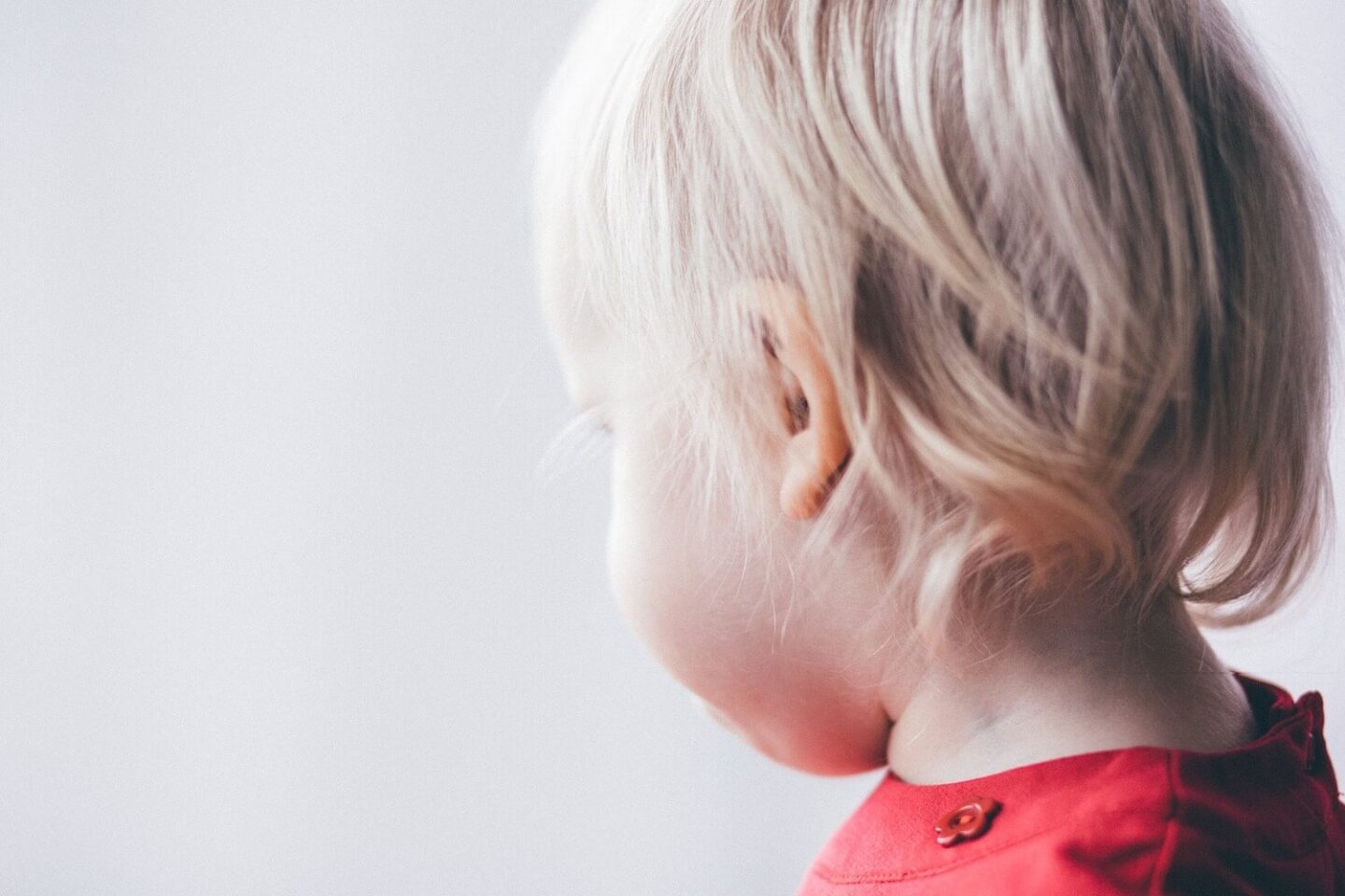 The side of a toddlers head. They have blonde hair and are wearing a red top