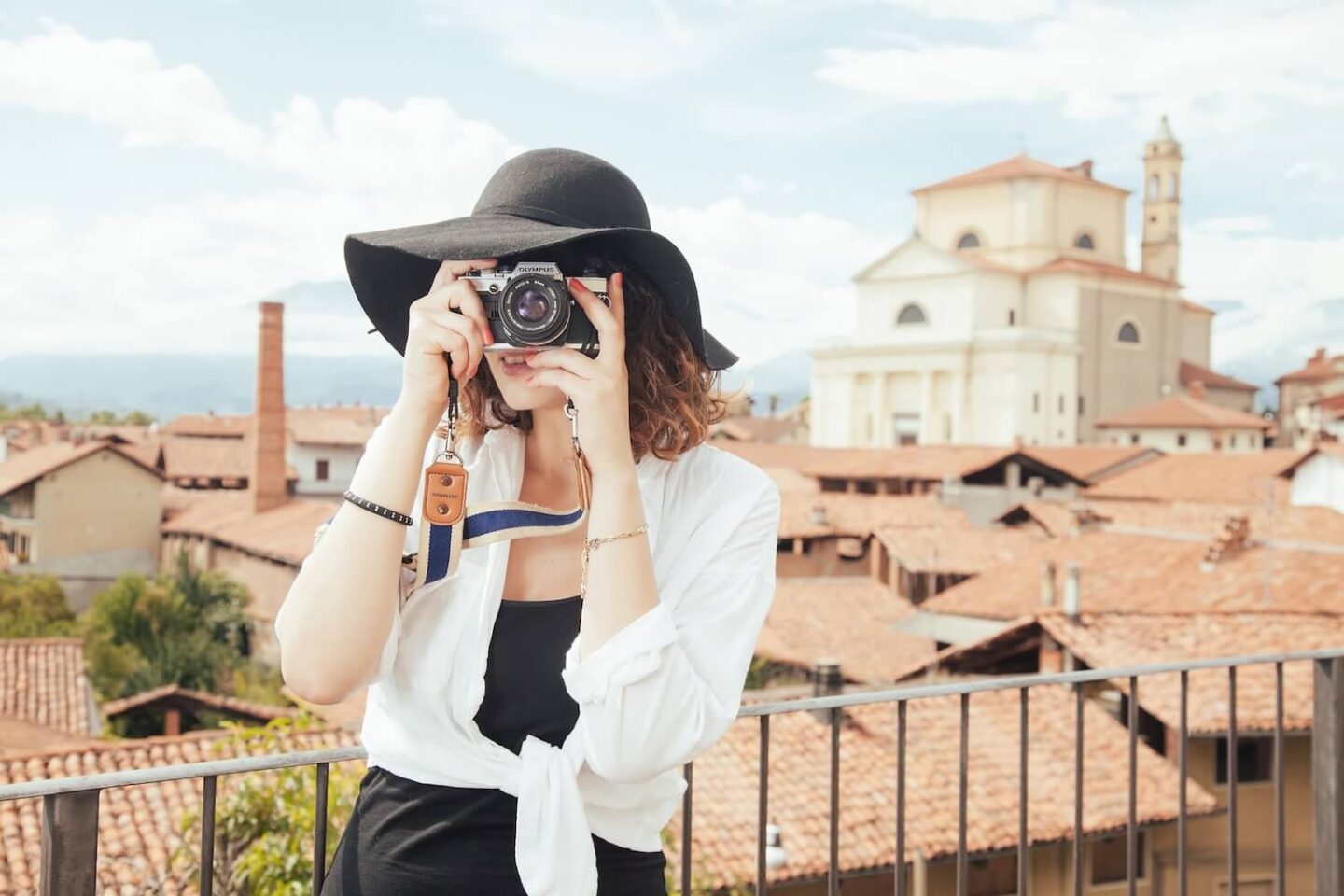 A woman standing on a balcony. She is holding a camera up to her eye as if she is taking a picture. Behind her is a view of rooftops