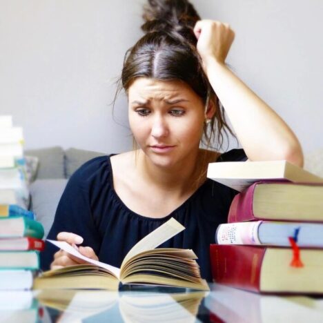 A woman sitting at a table with stacks of books. She looks frazzled as he looks at one of them