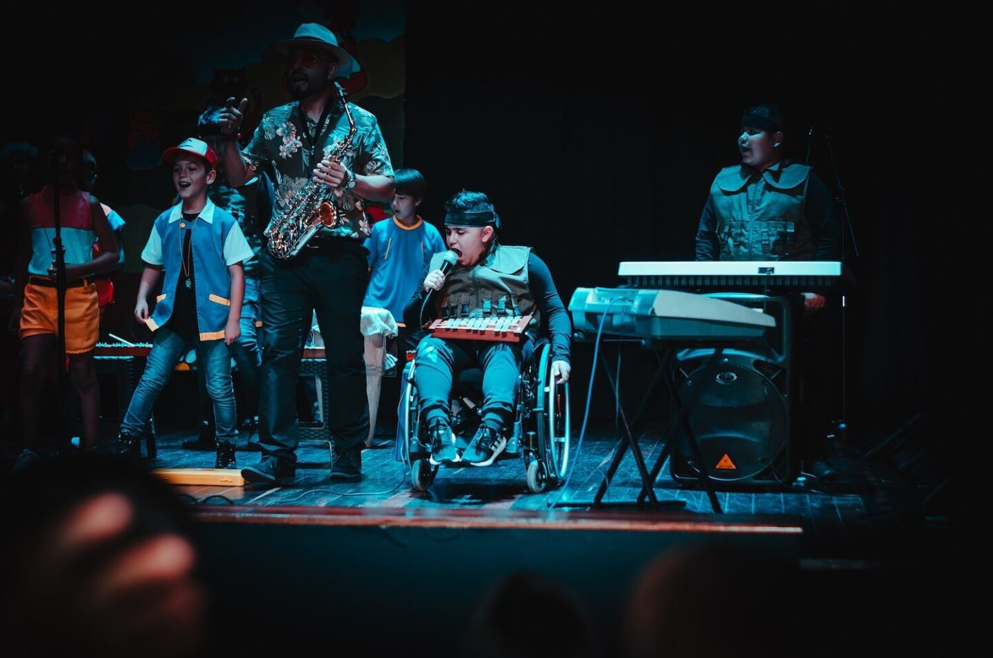 A group on a stage with musical instruments. A man with a saxophone appears to be leading and the rest of the band members are children.