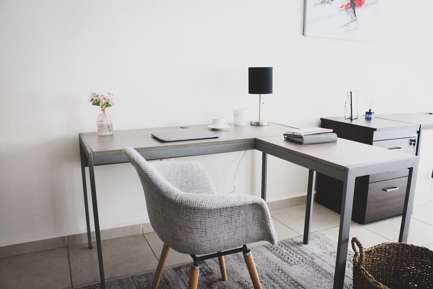 A corner desk with a lamp, laptop and little vase of flowers. A comfortable grey desk chair is in front.