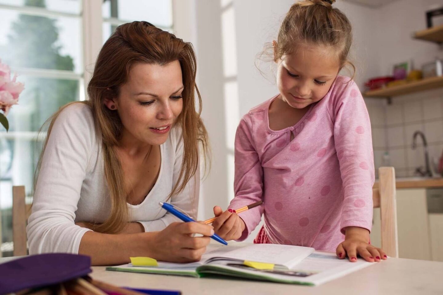 A woman and her daughter are sat at a table. They are looking down at some open workbooks and the woman has a pen in her hand which she is using to point at something on the book