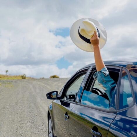 A blue car on a road in the countryside. An arm is coming out of the bar window holding a panama hat