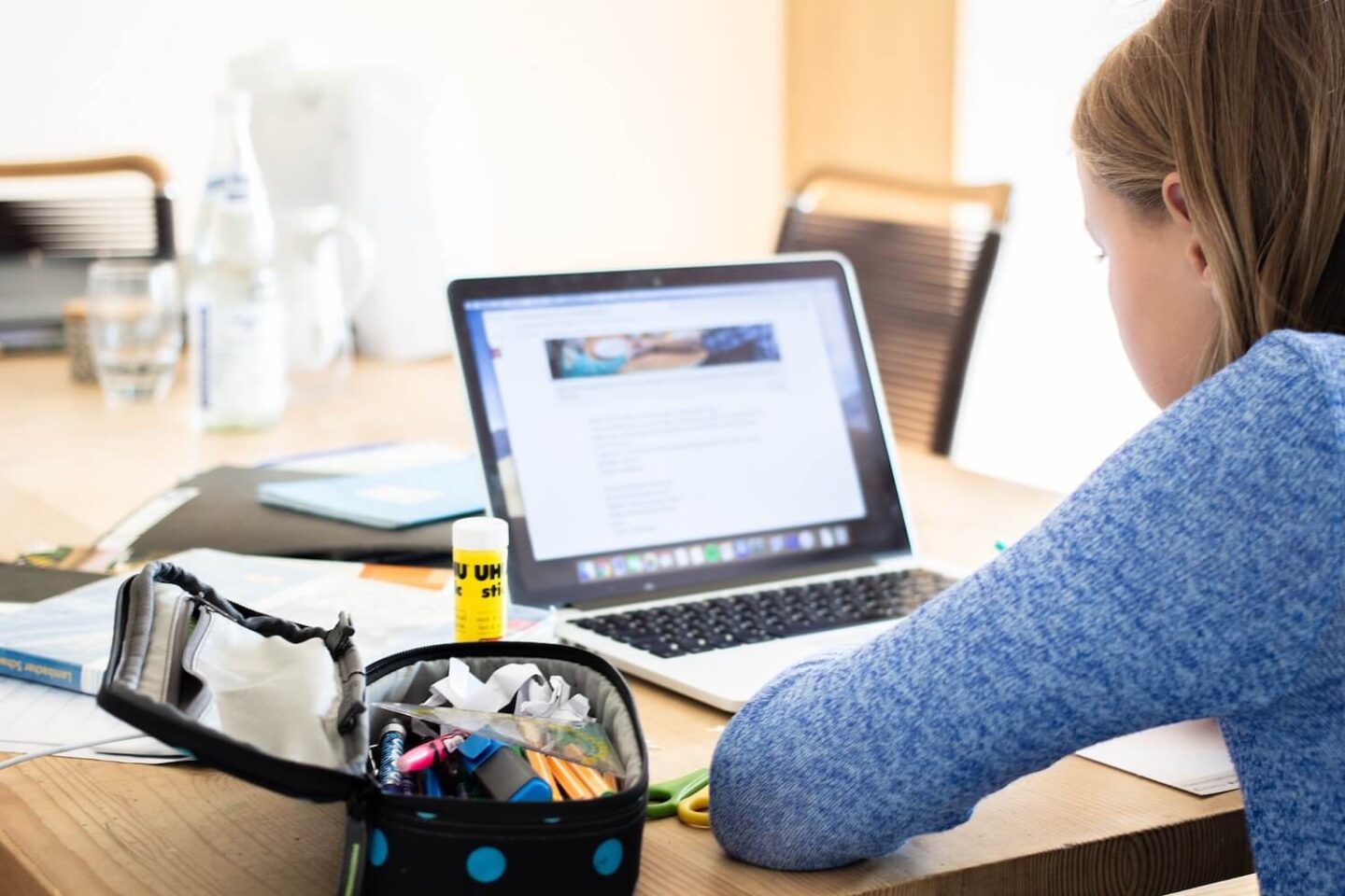A girl sitting and working on a laptop. She has a pencil case and glue stick with her.